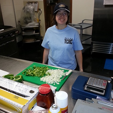 Woman in a TCRC shirt stands behind a cutting board of chopped onions and green peppers.